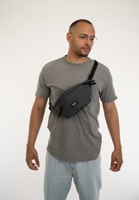 Borsa a tracolla nera in tessuto, indossata a bandoliera, con una tasca con zip, cinturino regolabile e etichetta con logo. Maglietta grigia casual e jeans.