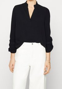 Blusa nera a maniche lunghe con scollo a V e spalle arricciate, con dettagli testurizzati sulle maniche. Abbinata a pantaloni bianchi.