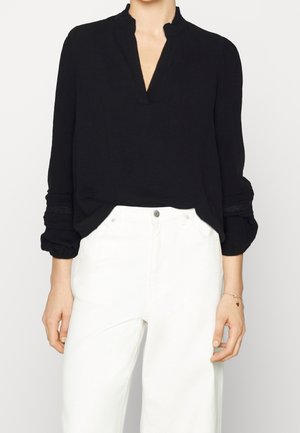 Blusa nera a maniche lunghe con scollo a V e spalle arricciate, con dettagli testurizzati sulle maniche. Abbinata a pantaloni bianchi.