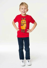 Rotes T-Shirt mit einem Comic-Charakter, der braune Ohren und einen orangefarbenen Körper hat; getragen mit dunkelblauen Hosen und weißen Turnschuhen.