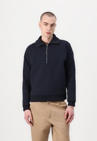Sudadera de peso medio en navy con cuello de media cremallera, puños y dobladillo acanalados. Textura suave, corte relajado y diseño minimalista.