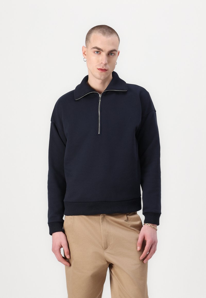 Sudadera de peso medio en navy con cuello de media cremallera, puños y dobladillo acanalados. Textura suave, corte relajado y diseño minimalista.