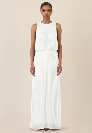 Frau trägt ein ärmelloses weißes, plissiertes Maxikleid und steht vor einem beigen Hintergrund mit entspannt an den Seiten hängenden Händen.