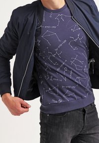 Giubbotto bomber blu sopra una maglietta blu con equazioni matematiche bianche e motivi geometrici. I jeans neri sono parzialmente visibili.