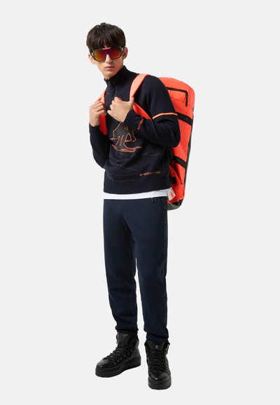 Marineblaue Fleecejacke mit orangen Akzenten, weißes T-Shirt, marineblaue Jogginghosen und schwarze Stiefel. Helle orangefarbene Rucksack mit schwarzen Streifen.