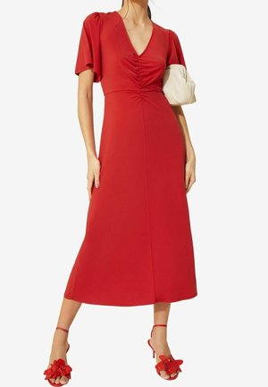 Femme portant une robe midi rouge à devant froncé, manches bouffantes, et sandales à talons hauts rouges à motif floral, tenant une pochette crème froncée.
