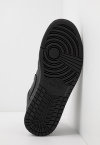 Semelle de basket Nike noire avec motifs de semelle circulaires et rectangulaires, placée à la verticale contre un mur blanc d’angle.