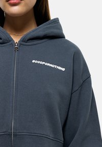 Felpa con cappuccio in tessuto blu scuro, con chiusura a zip e testo bianco "GOODFORNOTHING" sul petto. Tessuto morbido, taglio rilassato.