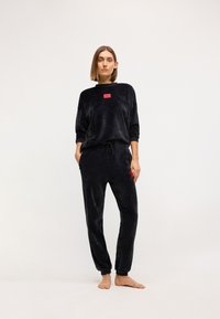 Čierny plyšový loungewear set obsahuje dlhý rukáv top s červenou logovou nášivkou a zodpovedajúce jogger nohavice s elastickým pásom a manžetami.
