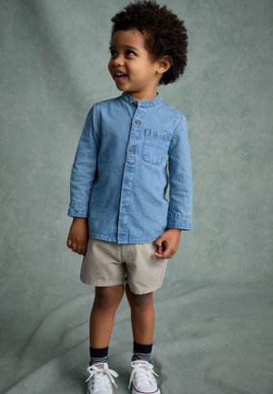 Junges Kind mit lockigem Haar, trägt ein blaues Hemd mit Knöpfen, beige Shorts, gestreifte Socken und weiße Turnschuhe, schaut nach rechts und lächelt.