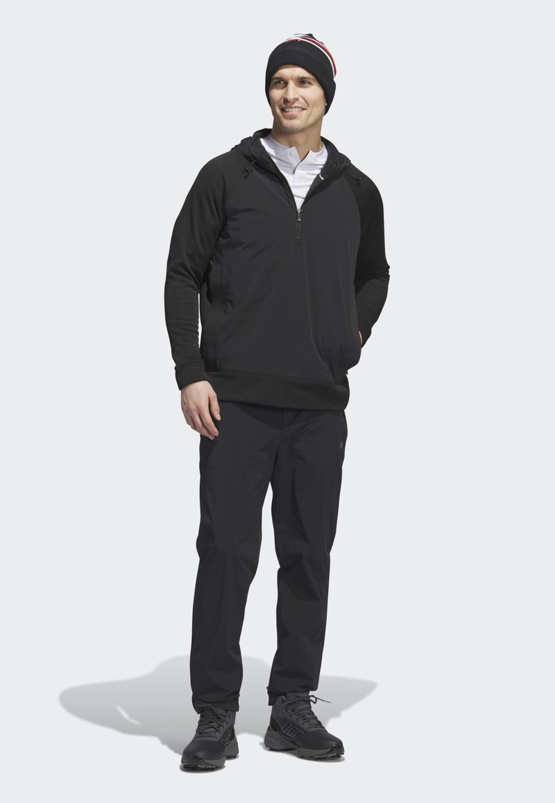 Schwarzer Hoodie mit Halb-Reißverschluss, passende schwarze Hose und schwarze Sneaker. Zubehör umfasst eine schwarze Strickmütze mit bunten Streifen.