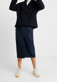 Marineblauwe dubbelgeknoopte jas met grote knopen, gecombineerd met een marineblauwe rok tot de knie met witte stiksels en zakken. Lichtgekleurde sneakers.