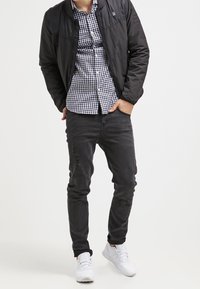 Giacca bomber nera sopra una camicia a quadri, abbinata a jeans neri strappati e scarpe da ginnastica bianche. Ensemble semplice e casual.