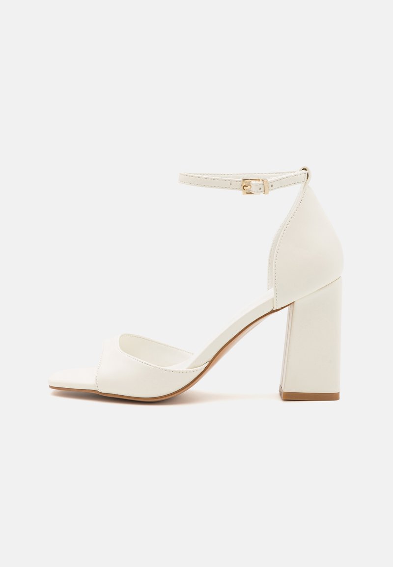 White high-heeled sandal with a block heel, open toe, ankle strap with gold buckle, and smooth synthetic material.