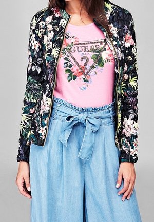 Woman wearing a floral quilted jacket, pink floral Guess logo t-shirt, and high-waisted light blue pants tied with a bow belt.