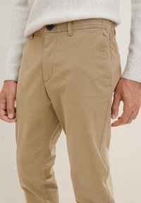 Beżowe spodnie typu chino o gładkiej fakturze, z zapinanym na guzik pasem i bocznymi kieszeniami, prezentowane na osobie noszącej jasny sweter.