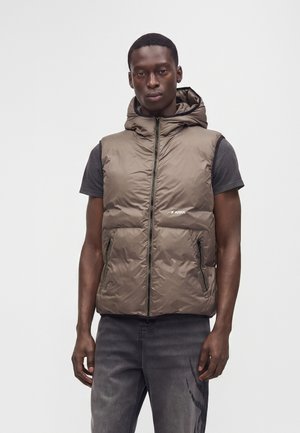 Homme debout portant une veste matelassée marron sans manches avec capuche par-dessus un t-shirt noir à manches courtes et un jean gris foncé sur un fond uni.