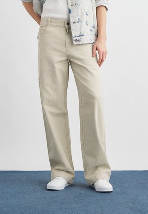 Person iført beige vid bukser med nålestriper, hvite joggesko, en hvit topp og en lyseblå denimjakke med oppbrettede ermer.