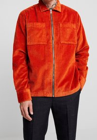 Giacca in corduroy arancione con collo, zip frontale e due tasche sul petto. Presenta un tessuto testurizzato e una vestibilità rilassata.