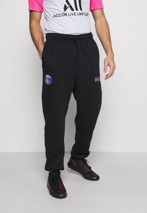 Mężczyzna noszący czarne dresy PSG z logo, białą i różową koszulkę Paris Saint-Germain oraz czarne sneakersy z czerwonymi akcentami.