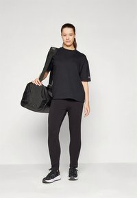 Črna bombažna majica z ohlapnim krojem, kombinirana s črnimi pajkicami. Model nosi črno športno torbo in črne športne copate.