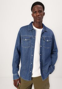 Denim srajca v srednje modri barvi z gumbi spredaj, dvema žepoma na prsih in klasičnim ovratnikom. Enostavna tekstura; oblečena čez belo majico.