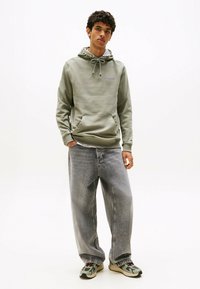 Giovane in piedi che indossa un hoodie verde chiaro, jeans grigi sbiaditi larghi e sneakers grigio-verdi su uno sfondo bianco.