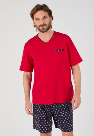 Man die een rood T-shirt met korte mouwen draagt met een patroon op de zak en donkerblauwe shorts met een kleine ankerprint op een effen achtergrond.