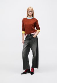 Maglione peloso rosso-arancione con scollo multicolore, abbinato a jeans grigi larghi e scarpe nere con punta, con calzini rossi e vestibilità rilassata.