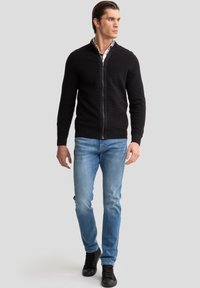 Sudadera negra con cremallera y diseño de punto texturizado, combinada con jeans azul claro y zapatillas negras. Modelo de pie contra un fondo neutro.