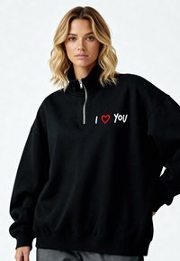 Sweat-shirt noir en polaire avec fermeture quart-zip et coupe ample. Présente un cœur rouge et le texte « JE T'AIME » en broderie blanche sur la poitrine. Texture douce.