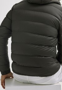 Dunkelgrüne Steppjacke mit Kapuze, ausgestattet mit gestepp-ten Bereichen und elastischen Bündchen. Wird über weißen Hosen getragen und zeigt eine elegante, glatte Textur.