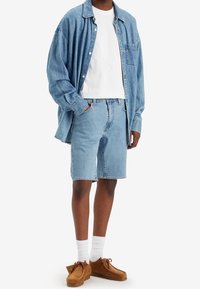 Camisa de mezclilla azul claro, camiseta blanca de cuello redondo, pantalones cortos de mezclilla a juego, zapatos de ante marrón y calcetines blancos. Look casual, de corte y diseño relajado.