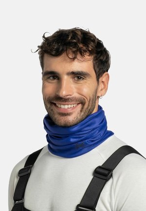 Smiling man with curly hair wearing a blue Buff neck gaiter, white shirt, and black overall straps against a plain background.