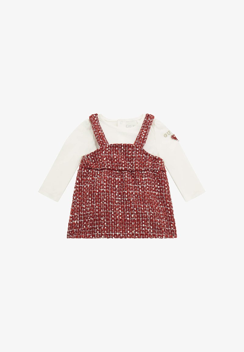 Red and white woven jumper dress with shoulder straps over a long-sleeve white top. Features a round neckline and textured fabric.