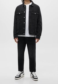 Personne portant une veste en denim noire par-dessus un t-shirt blanc, un jean noir et des baskets noir et blanc, debout sur un fond blanc.