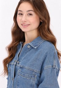 Ljusblå denimjacka med knappframsida, två bröstfickor och krage. Har en avslappnad passform och synliga sömnadsdetaljer.