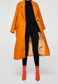 Abrigo largo y oversize de lana naranja con cuello muesca, que presenta un botón negro. Lleva puesto sobre una blusa negra y unos jeans, combinado con botas rojas de tobillo.