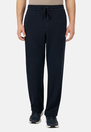 Mann trägt schwarze lockere Hose mit Kordelzug und schwarze Sneakers, steht vor weißem Hintergrund.