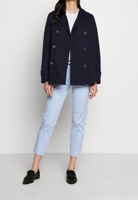 Marineblaue doppelte Jacke mit goldenen Knöpfen, getragen über einem weißen Hemd und blauen gestreiften Hosen, kombiniert mit schwarzen Slippern.