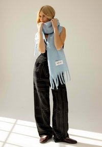 Écharpe bleu clair avec des franges drapée autour du cou, associée à un pantalon large noir avec des motifs géométriques blancs. Texture douce et confortable.