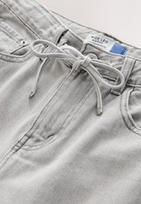 Jean gris clair à jambes larges, taille mi-haute, avec poches avant et un cordon noué en nœud à la ceinture.