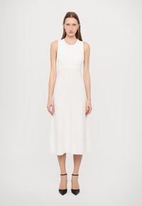 Abito bianco senza maniche con corpetto aderente e gonna midi svasata. Tessuto a maglia liscia e design minimal. Indossato con tacchi neri con cinturino alla caviglia.