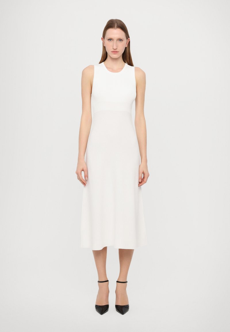 Abito bianco senza maniche con corpetto aderente e gonna midi svasata. Tessuto a maglia liscia e design minimal. Indossato con tacchi neri con cinturino alla caviglia.