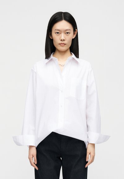 Jeune femme aux cheveux noirs raides porte une chemise blanche surdimensionnée boutonnée et un pantalon noir, debout devant un fond uni clair.