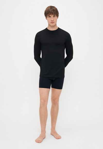 Juodos ilgomis rankovėmis viršus su prigludusia forma, derinamas su tamsiomis šortais. Audinys atrodo lygus ir lengvas. Matomų raštų ar akcentų nėra.