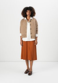 Beige strikket cardigan med knappelukking, lagdelt over en hvit skjorte, kombinert med et oransje skjørt og mørke loafers med gullaccent.