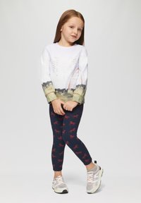 Chemise blanche à manches longues avec un imprimé paysage et un arc-en-ciel, assortie à un legging bleu foncé orné de motifs de chevaux rouges. Baskets grises.