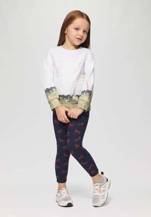Weißes Langarmhemd mit Landschaftsprint und Regenbogen, kombiniert mit dunkelblauen Leggings mit roten Pferdemustern. Graue Sneaker.
