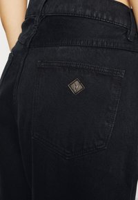 Svarta denimjeans med en bakficka som har en diamantformad lapp i brunt läder. Tyget har en lätt struktur och matt yta.